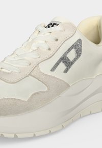 Gros plan d'une sneaker blanche et beige avec des détails en suède, des lacets blancs, une semelle épaisse et un logo argenté scintillant sur le côté.
