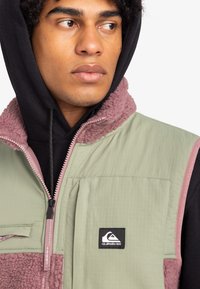 Grünes Westchen mit einem plüschigen rosa Kragen und einem schwarzen Kapuzenpullover darunter. Verfügt über einen Reißverschluss und ein Logo-Patch auf der Brusttasche.