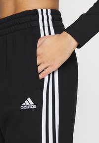 Ruka v kapse černých kalhot Adidas s bílými pruhy a logem, nošena s černým dlouhým rukávem, zkráceným na úroveň pasu.
