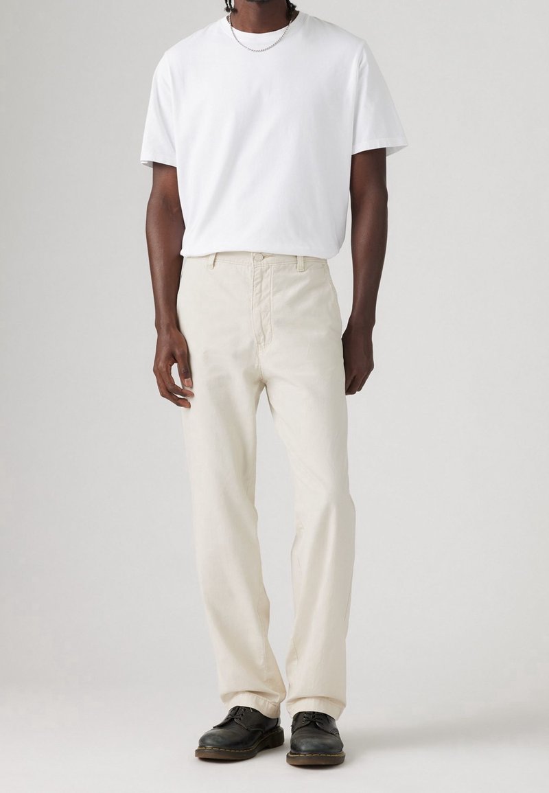 Homme portant un t-shirt blanc uni, un pantalon couleur crème, des chaussures noires et un collier chaîne en argent, debout devant un fond clair uni.