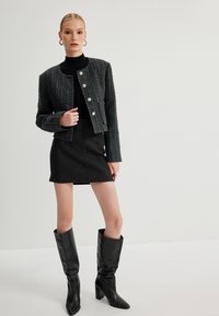 Gestructureerde donkergrauwe tweed cropped jack met zilveren knopen, gecombineerd met een strakke donkere rok en kniehoge zwarte leren laarzen.