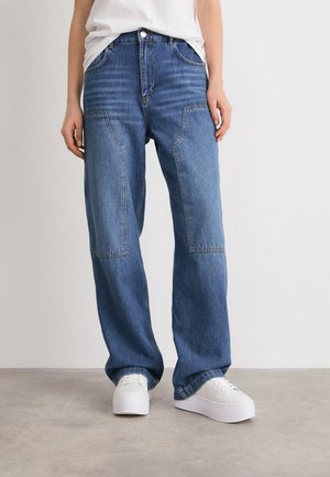 Personne portant un jean bleu ample avec de grandes poches plaquées à l'avant et des baskets blanches à plateforme, debout sur un sol gris contre un mur blanc.