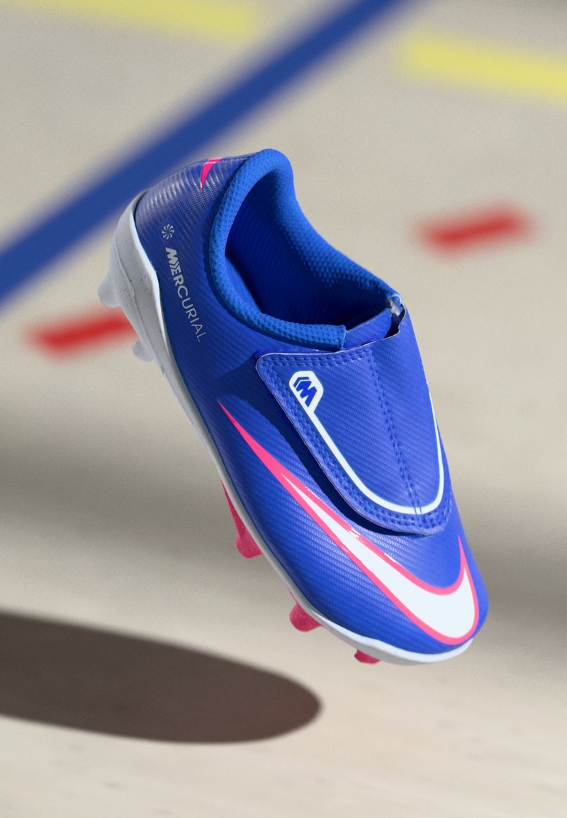 Blå Nike Mercurial fodboldstøvle med lyserøde accenter og såler, svævende over et sportsbanegulv med farvede linjer.