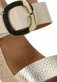 Sandalia de plataforma de cuero beige con una amplia tira metálica dorada y un hebilla cuadrada. La parte superior tiene un acabado texturizado y la suela es color marrón claro.