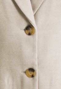 Beige stof blazer met een gestructureerd ontwerp, twee grote ronde knopen met een schildpadpatroon en een gladde, textuurachtige afwerking.