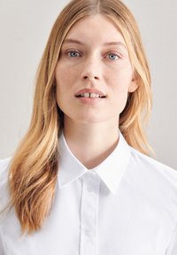 Camicia bianca con collare a punta, caratterizzata da una texture liscia e una leggera lucentezza, indossata da una persona con capelli lunghi e lisci.