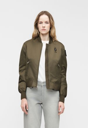 Femme portant une veste bomber vert olive sur un t-shirt blanc et un jean gris clair, debout devant un fond clair uni.