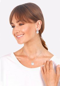 Orecchini circolari in tonalità argento con superfici testurizzate, abbinati a una collana con pendente coordinato su un indumento bianco, evidenziando un design minimalista.