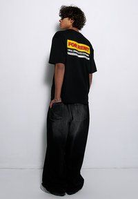 Czarny, oversize t-shirt z wyraźnym żółtym i czerwonym napisem "DO WYNAJĘCIA" na plecach. Połączony ze luźnymi czarnymi dżinsami.