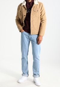 Giacca di jeans beige con collo in shearling, felpa bordeaux sotto, jeans azzurri e sneaker bianche in posizione eretta.