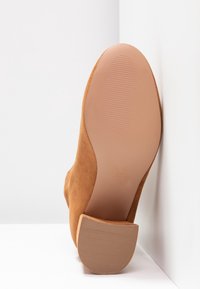 Bottine en daim couleur tan avec une semelle texturée en caoutchouc et un talon empilé, vue du dessous, mettant en valeur des surfaces lisses et rainurées.