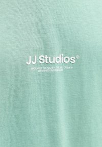 Tissu vert clair avec texte blanc "JJ Studios" et texte plus petit "Présenté par The J&J Crew, conçu au Danemark."