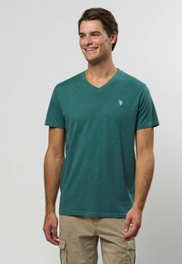 Grön v-ringad t-shirt i mjukt material, med korta ärmar och en liten broderad logotyp på vänster bröst. Neutral bakgrund.