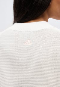 Κοντινό πλάνο ενός ατόμου που φοράει ένα λευκό πλεκτό πουλόβερ με ένα μικρό, ανοιχτό ροζ λογότυπο Adidas κεντημένο στο πάνω μέρος της πλάτης.