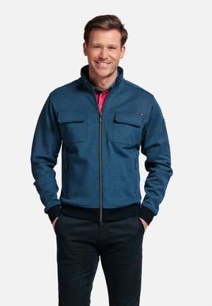 Blauwe zip-up jas met hoge kraag, twee borstzakken en elastische manchetten. Draagt over een roze polo. Lichtgewicht stof.