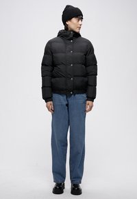 Černá prošívaná bunda s přiléhavým střihem, zapínáním na cvoky a vysokým límcem, v kombinaci s volnými modrými džínami a černými botami.