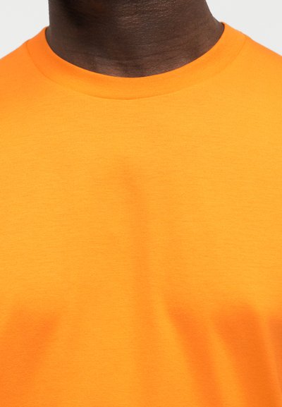 Κοντινό πλάνο ενός ατόμου που φοράει μια μονόχρωμη φωτεινή πορτοκαλί μπλούζα με στρογγυλή λαιμόκοψη, δείχνοντας τον λαιμό και το άνω μέρος του στήθους.