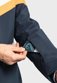 Marineblaue wasserdichte Jacke mit einem orangefarbenen Streifen, ausgestattet mit einer Reißverschlusstasche am Ärmel. Eine Hand steckt eine Karte in die Tasche.