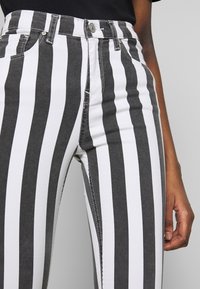 Primer plano de una persona que lleva pantalones de rayas verticales en blanco y negro con botón y cremallera, con la mano relajada al costado.