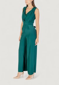 Una mujer vestida con un mono sin mangas de color verde profundo se muestra con confianza en tacones nude contra un fondo claro.