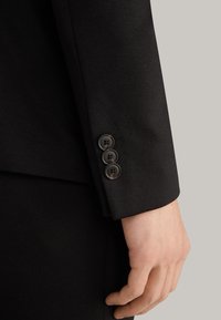 Schwarze maßgeschneiderte Anzugjacke mit Ärmeln und drei dunklen Knöpfen sowie subtilen Nähdetails. Der Stoff wirkt glatt mit einem matten Finish.