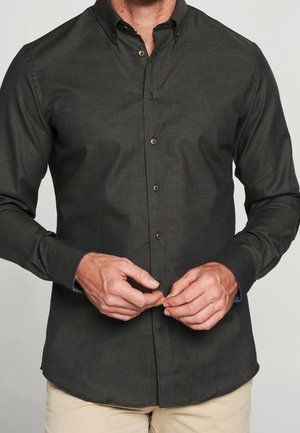 Mies, joka pukeutunut tummanharmaaseen napitettuun pitkähihainen paitaan, napin kiinnitys, yhdistetty beigestä housuihin yksivärisestä taustasta.