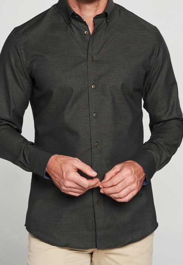 Homme portant une chemise à manches longues gris foncé boutonnée, en train de boutonner un bouton, associée à un pantalon beige sur un fond uni.