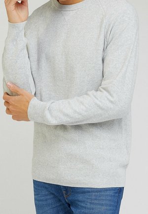 Muž v lehkém šedém pleteném svetru s texturou a modrých džínách, upravující rukáv jednou rukou na jednotném pozadí.