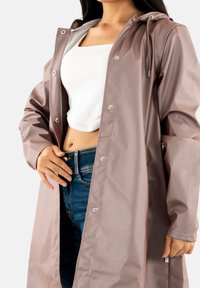 Raincoat waterproof color lilla chiaro, realizzato in materiale lucido, con cappuccio, bottoni a pressione e vestibilità comoda, abbinato a jeans blu.