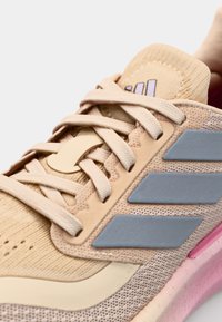 Beige athletic shoe with mesh upper, light gray accents, and purple branding. Features traditional laces and textured sole.