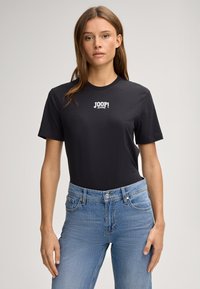 Schwarzes Baumwoll-T-Shirt mit kurzen Ärmeln und rundem Halsausschnitt, mit weißem "JOOP! JEANS" Logo auf der Vorderseite. Kombiniert mit hellblauen Jeans.