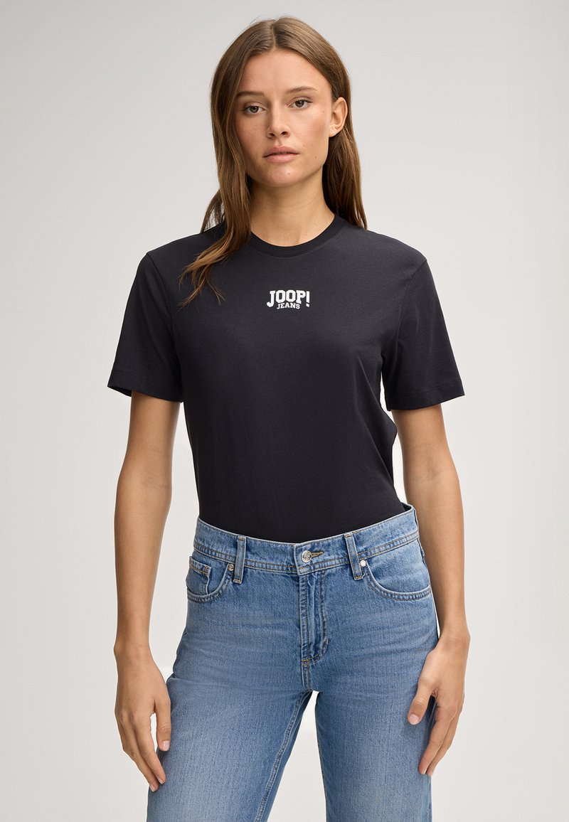Schwarzes Baumwoll-T-Shirt mit kurzen Ärmeln und rundem Halsausschnitt, mit weißem "JOOP! JEANS" Logo auf der Vorderseite. Kombiniert mit hellblauen Jeans.