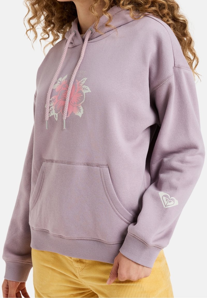 Personne aux cheveux bouclés portant un sweat à capuche lilas avec un motif floral rose et un logo blanc sur la manche, associé à un pantalon en velours côtelé jaune.
