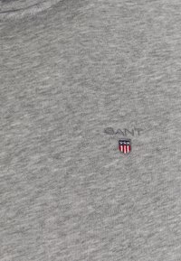 Graues Baumwoll-T-Shirt mit Rundhalsausschnitt, subtiler Textur und gesticktem "GANT"-Logo mit einem Schilddesign in Rot, Weiß und Blau.