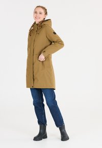 Brun, lång parkas med huva, dragkedja och knappstängning, sidofickor och elastiska muddar; bärs med blå jeans och svarta gummistövlar.