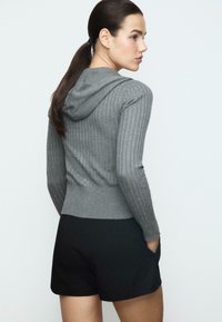 Donna con capelli scuri raccolti in una coda di cavallo, indossa un hoodie grigio a coste e pantaloni corti neri, in piedi con le mani in tasca, rivolta a sinistra su uno sfondo neutro.