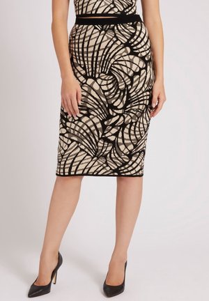 Femme portant une jupe beige longueur genou avec un motif abstrait noir et des talons hauts noirs, debout devant un fond uni.