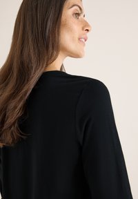 Musta vaate, jossa on sovitettu muoto ja pitkät hihat, valmistettu pehmeästä kankaasta. Hiukset valuvat yhden olkapään yli, korostaen kaula-aukkoa ja olkasaumaa.