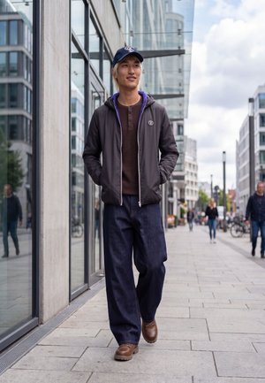 Un jeune homme portant une casquette, une veste, un jean et des bottes marche sur un trottoir en ville, passant devant des bâtiments modernes aux façades vitrées.