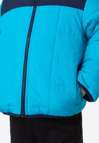 Niebieska pikowana kurtka puchowa z jednolitym granatowym górnym panelem, zamkiem błyskawicznym i elastycznymi mankietami, noszona na czarnych spodniach.