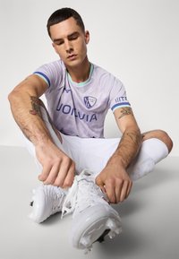 Un homme portant un maillot de football VfL Bochum violet clair et un short blanc est assis par terre en train de lacer des chaussures de football blanches. Des tatouages sont visibles sur ses bras et il porte des chaussettes blanches.