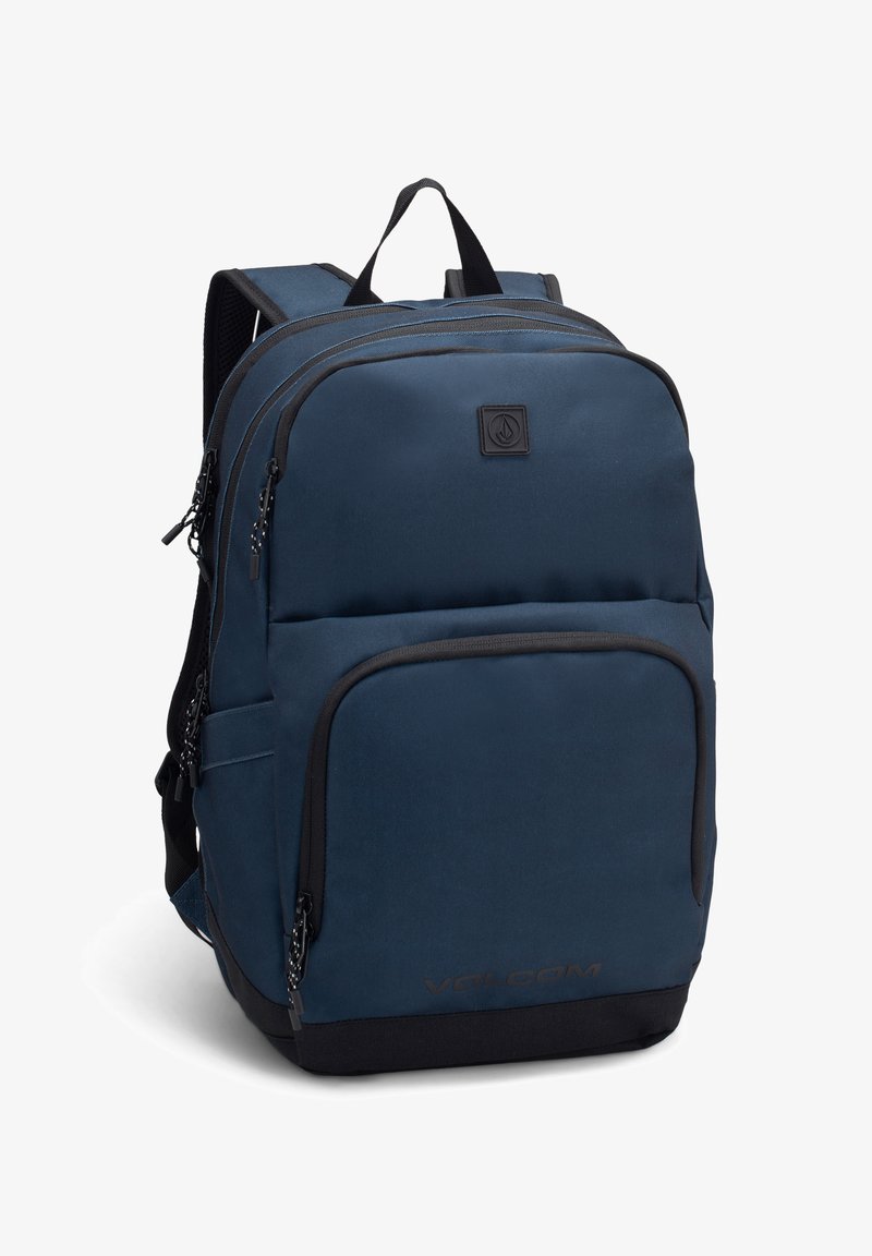 Granatowy plecak wykonany z trwałego materiału, z dwiema zamykanymi na zamek kieszeniami, wyściełanymi paskami oraz naszywką z logo z przodu.