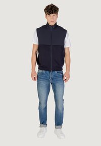 Gilet blu navy con zip da uomo, con accenti azzurri, abbinato a jeans chiari e sneaker bianche; presenta un colletto a righe e tasche laterali.