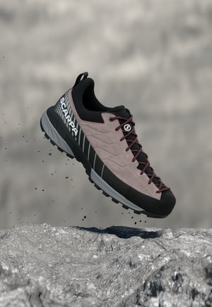 Svetlo vijolični in črni pohodni čevlji z bordo rdečimi vezalkami in logotipom SCARPA, v zraku nad kamnito površino z delci prahu okoli.