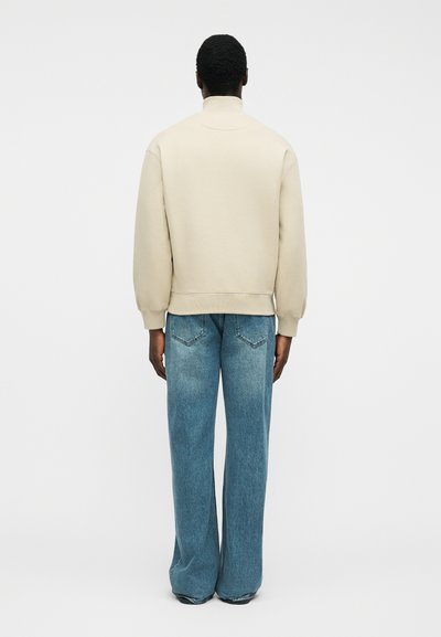 Άτομο που φοράει μπεζ μακρυμάνικο φούτερ και μπλε τζιν με φαρδιά μπατζάκια, στέκεται με την πλάτη προς τα εμπρός σε απλό λευκό φόντο.