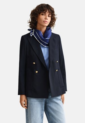 Donna con capelli ricci indossa un blazer doppiopetto blu navy con bottoni dorati, camicia azzurro chiaro, sciarpa blu e jeans chiari su sfondo bianco.