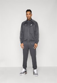 Grå træningsdragt lavet af glat stof, med en lynlåsjakke og matchende bukser med hvide sidestropper. Hvide og sorte sneakers med mesh.
