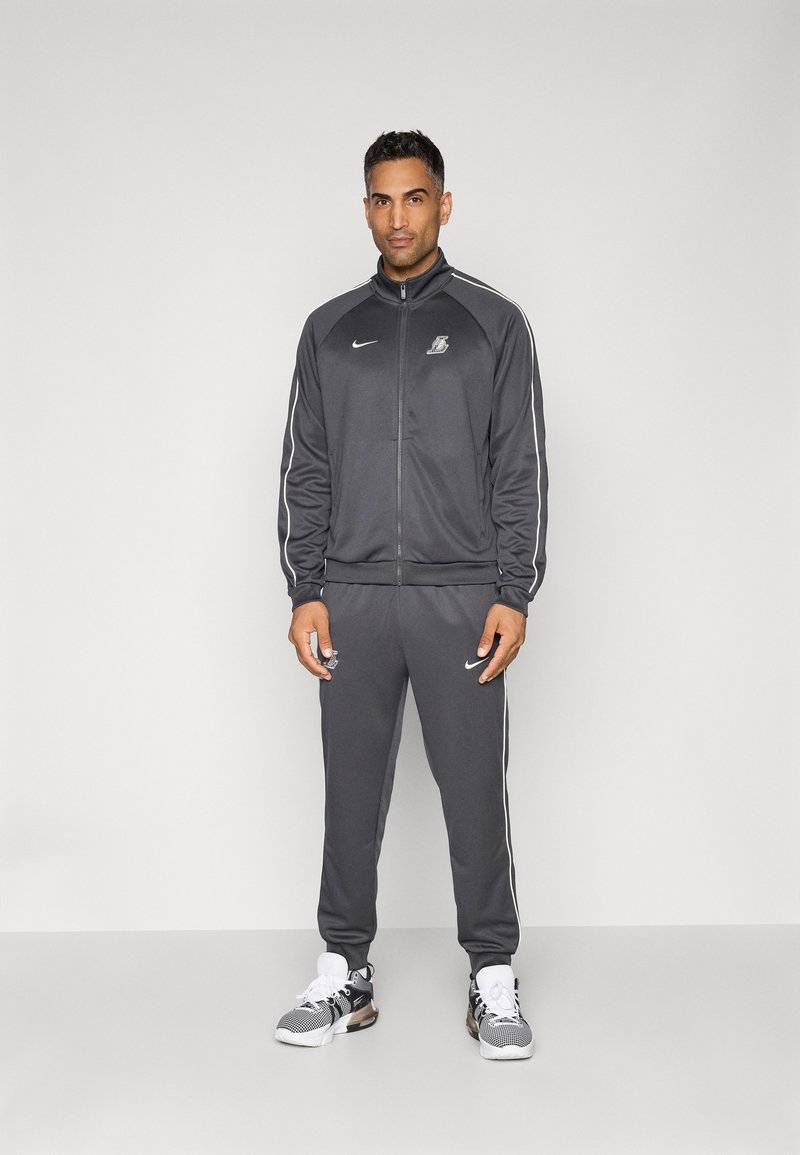 Grå træningsdragt lavet af glat stof, med en lynlåsjakke og matchende bukser med hvide sidestropper. Hvide og sorte sneakers med mesh.