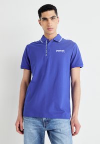 Fialová polo košeľa s bielym golierom a logom na ľavej strane. Krátke rukávy, bavlnený materiál, hladká textúra, príjemný strih.