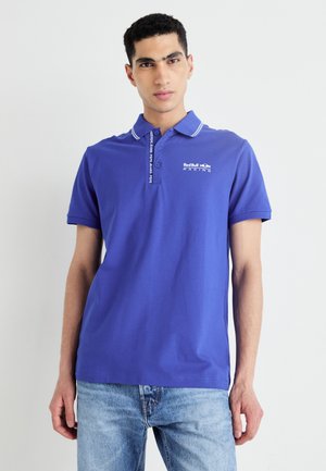 Lila polo-shirt med vit krage och logotyp på vänster sida. Kort ärm, bomulls-material, slät textur, avslappnad passform.